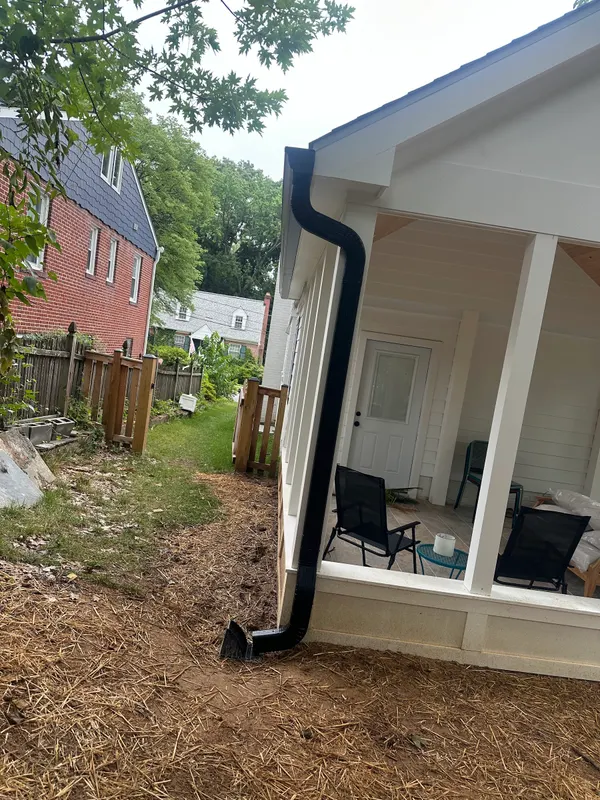 A black downspout running from a new gutter down the corner of a white porch and out onto a mulched bed.